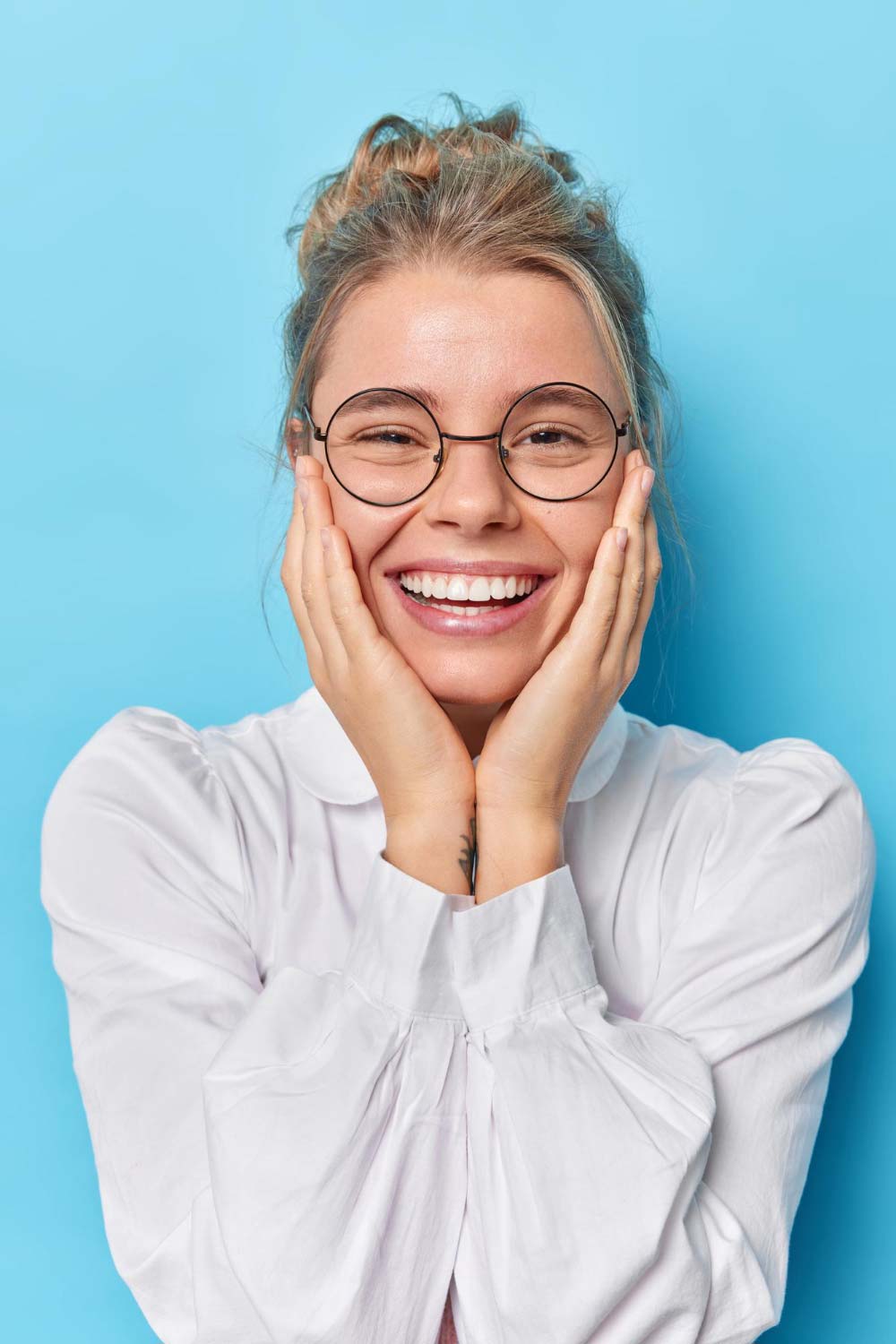 TPD-good-looking-lovely-european-woman-keeps-both-hands-cheeks-smiles-broadly-dressed-white-shirt-transparent-eyeglasses-hears-pleasant-news-poses-against-blue-background-looks-cute-camera